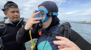 Dr. Mona Minkara has full scuba gear on with goggles over her face. She stands next to her Filipino scuba instructor Arnel. The background is the ocean and a blue sky.