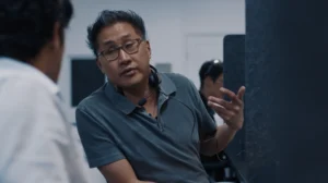A man with tan skin, black hair combed up, glasses, and gray shirt looks over at someone. He also has headphones with a mic around his neck and is in a cubicle office environment.