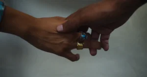 Two hands that are brown have their fingers gently intwined, but not fully grasping each other. One hand has rings on.