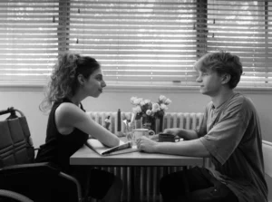 A black and white image of a young woman in a wheelchair with her hair pulled back in a curly ponytail in a sleeveless black dress. She sits at a restaurant table looking at the young man opposite her with a loose T-Shirt. There are flowers on the table and the background is blinds.