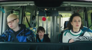 Three people sit in an accessible van with the grates of a folded ramp behind them. The driver is a woman with a sweater and a zebra steering wheel. In the passenger seat is a bored teenager with glasses and a blue hoodie. Behind them is a kid in a wheelchair.
