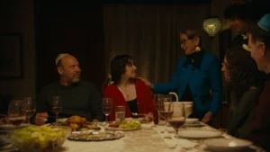 A darkly lit scene of a Passover Seder. There is a large table with a beige table cloth and plates of food. Noa, a young adult, wears a red sweater and sits next to her father. Noa looks up at her Grandma who has a hand on her shoulder, wearing a blue sweater. Three other people at the right of the frame look over at Noa.