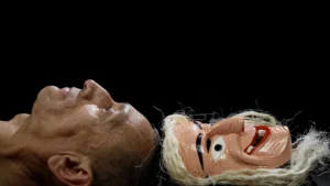 Against a black background there are two faces lying flat, the crowns of their heads touching. The face on the left is Ananias, a Mexican man who stares solemnly up. The face on the right is a mask with white hair, red lips, and two teeth. 