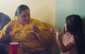 A Mexican woman wears a yellow sweater and is in the middle of signing, with her hands up to her chest. She stares off to the side at someone next to her off camera. Behind her is a white wall, and in front of her is a table with a red solo cup and a phone.